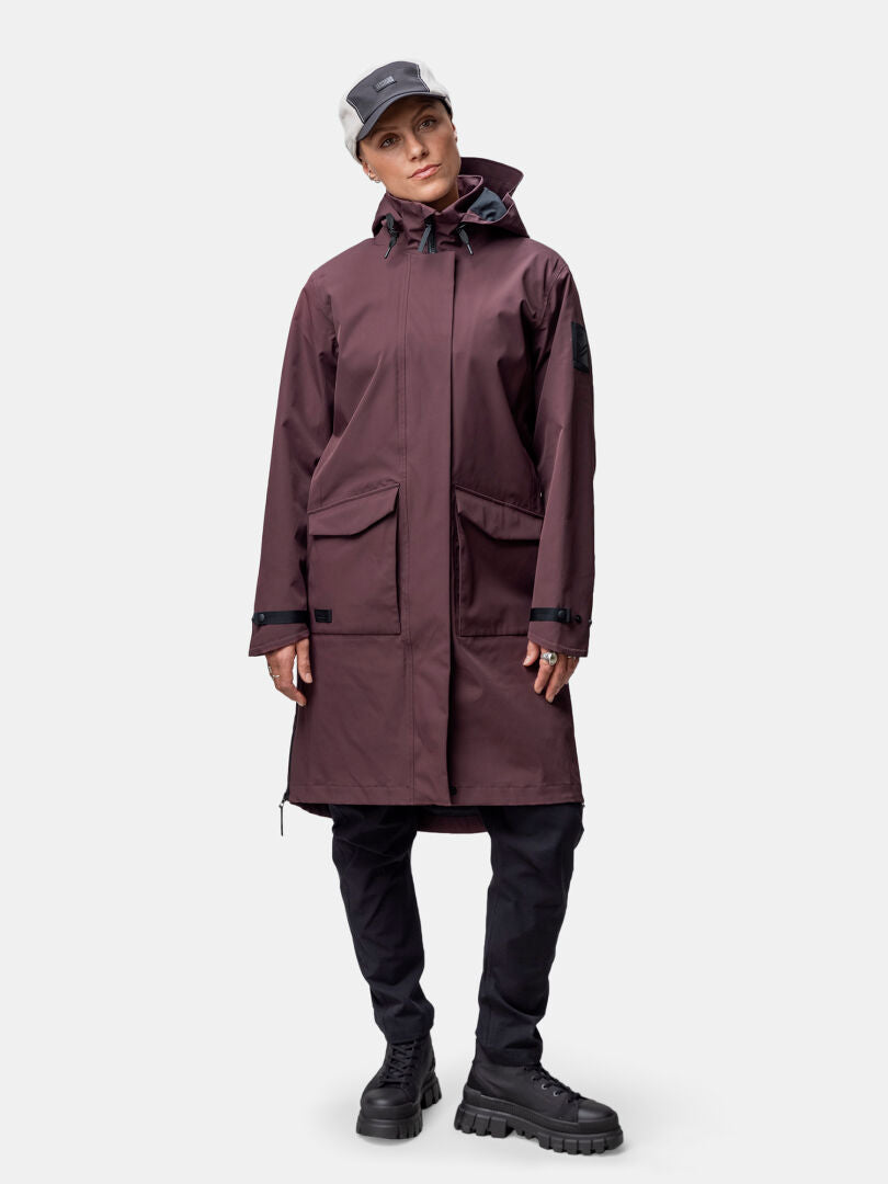 Eine Person in einer kastanienbraunen Halti Tokoi II DX Parkajacke Damen steht vor einem einfarbigen Hintergrund.