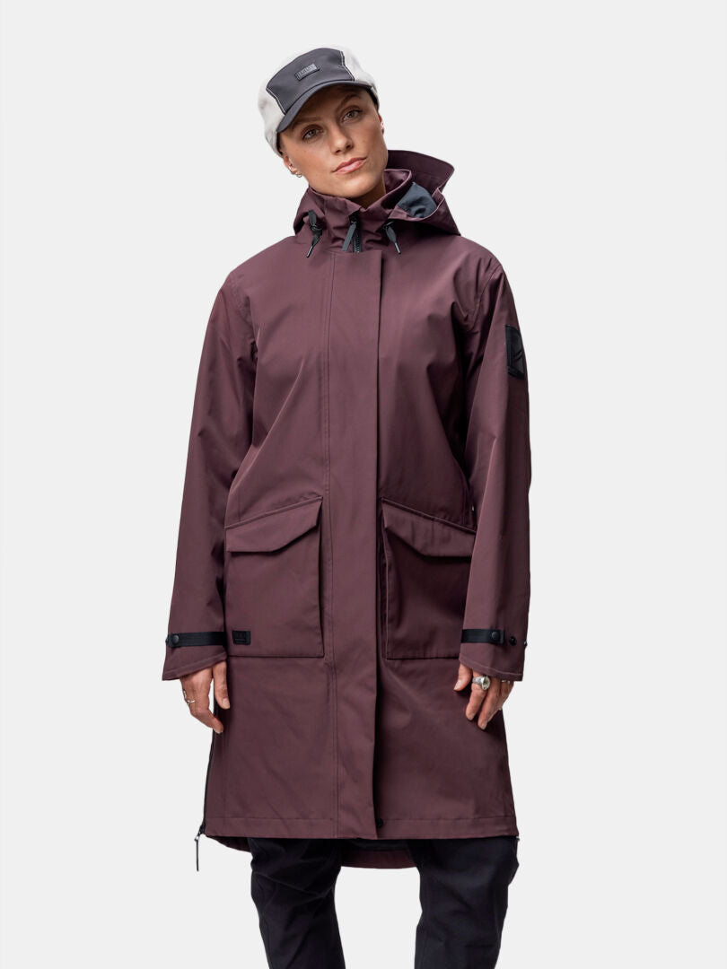 Eine Frau in einer kastanienbraunen Halti Tokoi II DX Parkajacke Damen steht vor einem schlichten weißen Hintergrund.