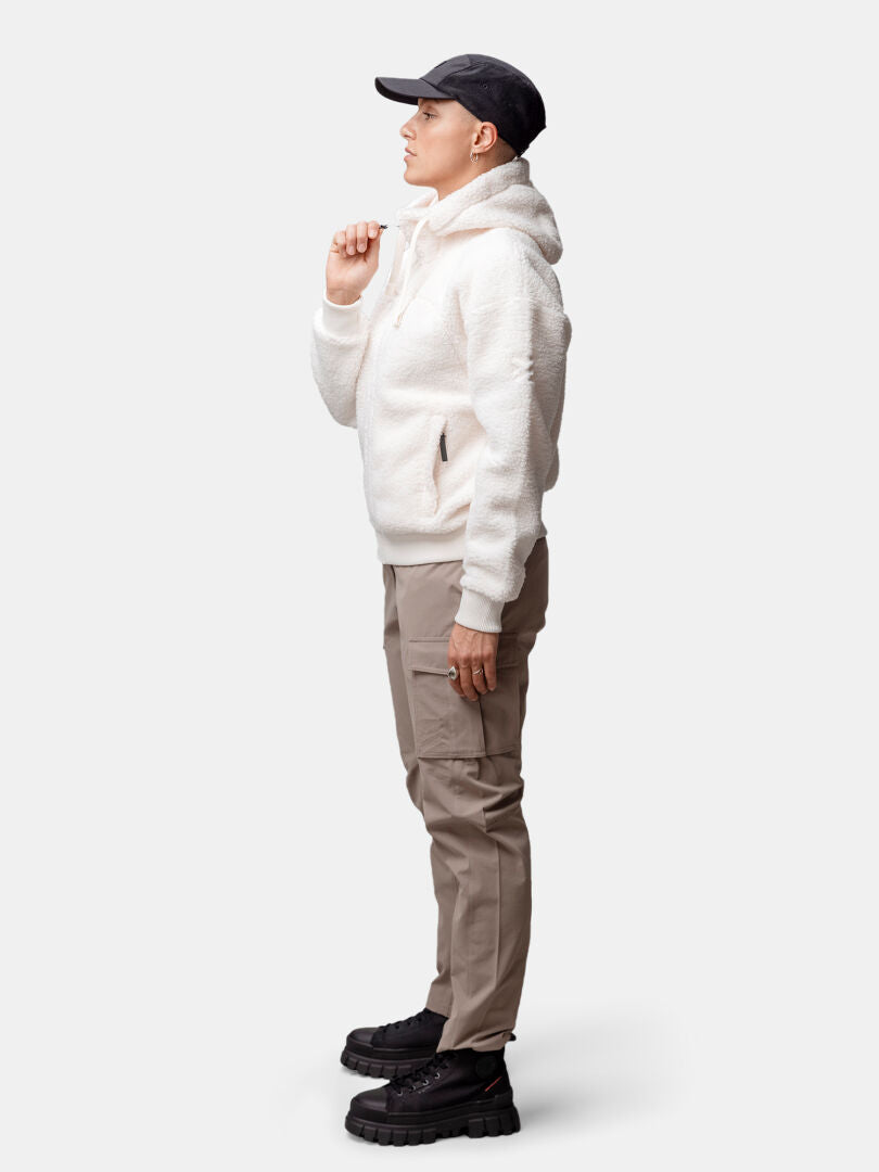 Eine Person in einer weißen Halti Lykky Fleece Jacke Damen, einer beigen Hose und einer schwarzen Mütze steht im Profil vor einem schlichten Hintergrund.