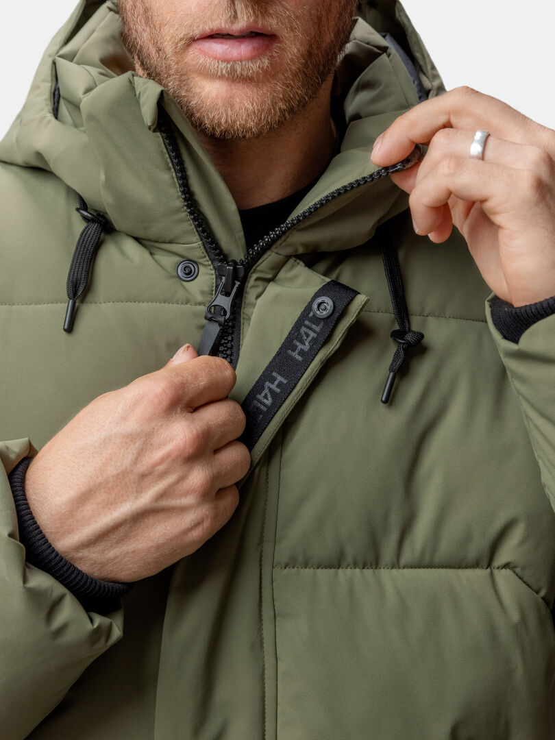 Ein Mann schließt den Reißverschluss der olivgrünen Brahe Puffer Winterjacke Herren von Halti, wobei das Logoschild und der silberne Ringzug zum Vorschein kommen.