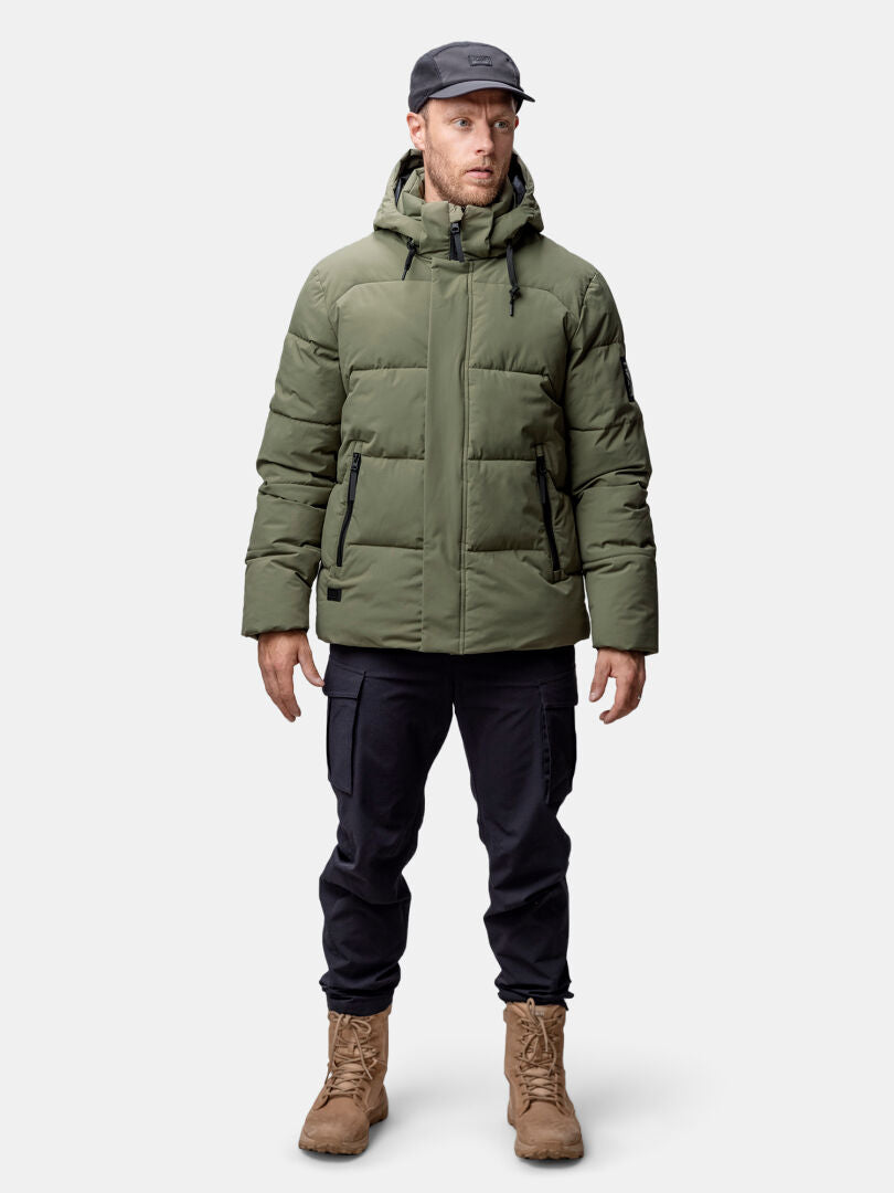 Ein Mann sieht nach vorne und trägt eine Halti Brahe Puffer Winterjacke Herren, eine schwarze Hose und hellbraune Stiefel.