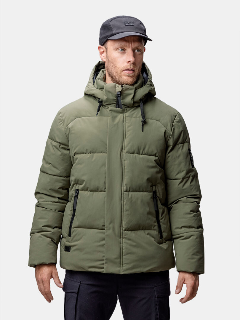 Ein Mann in einer urbanen olivfarbenen Halti Brahe Puffer Winterjacke Herren aus recyceltem Polyester steht vor einem schlichten hellen Hintergrund.