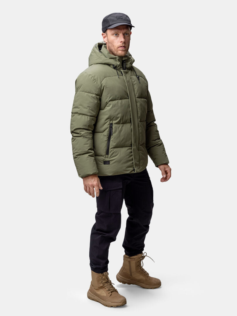 Ein Mann trägt eine Halti Brahe Puffer Winterjacke Herren in Olivgrün, eine schwarze Hose und hellbraune Stiefel.