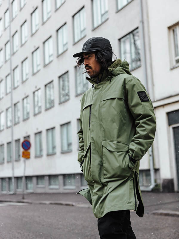 Eine Person in einer Halti Tokoi II DX Parka Jacke Herren und schwarzer Mütze geht auf einer Stadtstraße an einem Gebäude mit Fenstern vorbei.