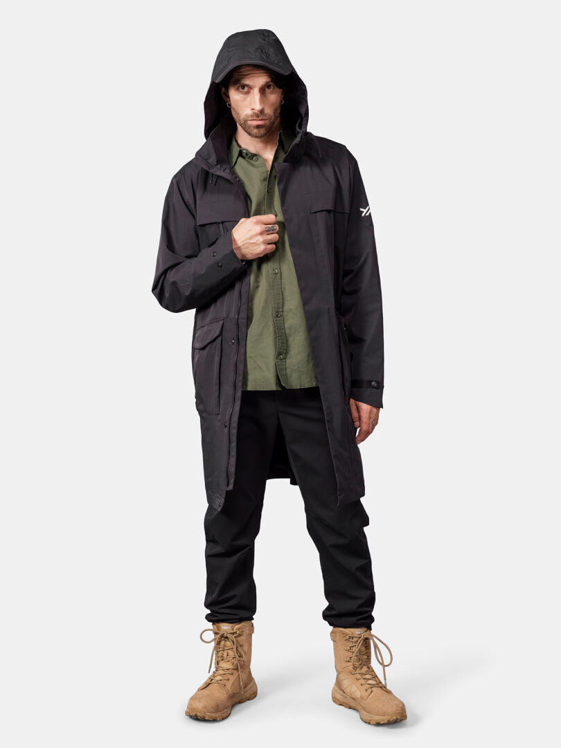 Ein Mann in einem „Halti Tokoi II DX“-Parka, einem grünen Shirt, einer schwarzen Hose und hellbraunen Stiefeln vor einem urbanen, hellen Hintergrund.