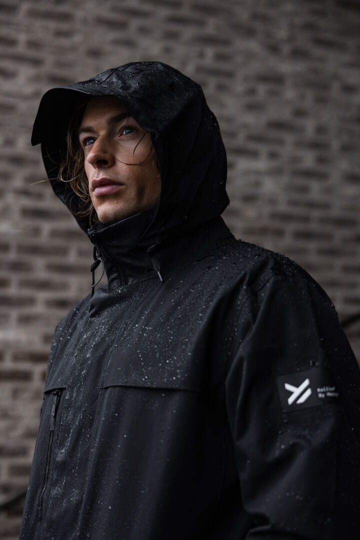 Ein Mann in einer schwarzen Halti Tokoi II DX Parka-Jacke mit Kapuze steht an einer Backsteinmauer, während Regentropfen an der Jacke abperlen.