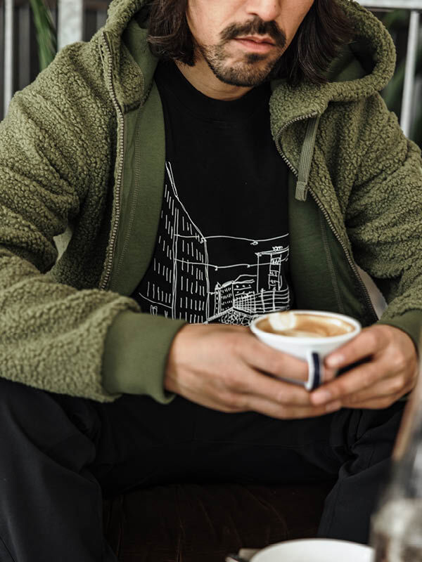 Mann in grüner Halti Lykky Fleece Jacke Herren, mit Kaffee in der Hand, schwarzes Hemd mit weißem Stadtbildmotiv.
