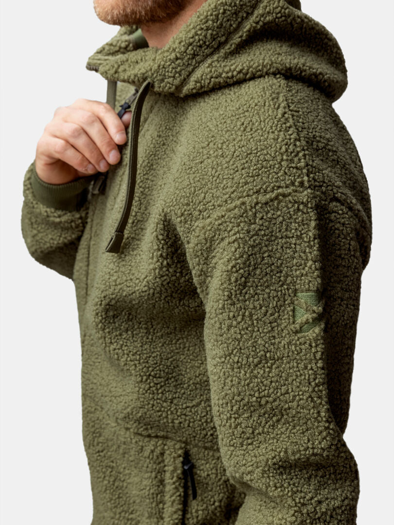 Eine Person trägt eine Halti Lykky Fleece Jacke Herren in Olivgrün mit einer Reißverschlusstasche, hergestellt aus recyceltem Polyester.