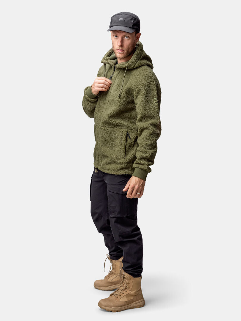 Mann in Halti Lykky Fleece Jacke Herren (oliv), schwarzer Cargohose und hellbraunen Stiefeln vor einfarbigem Hintergrund.