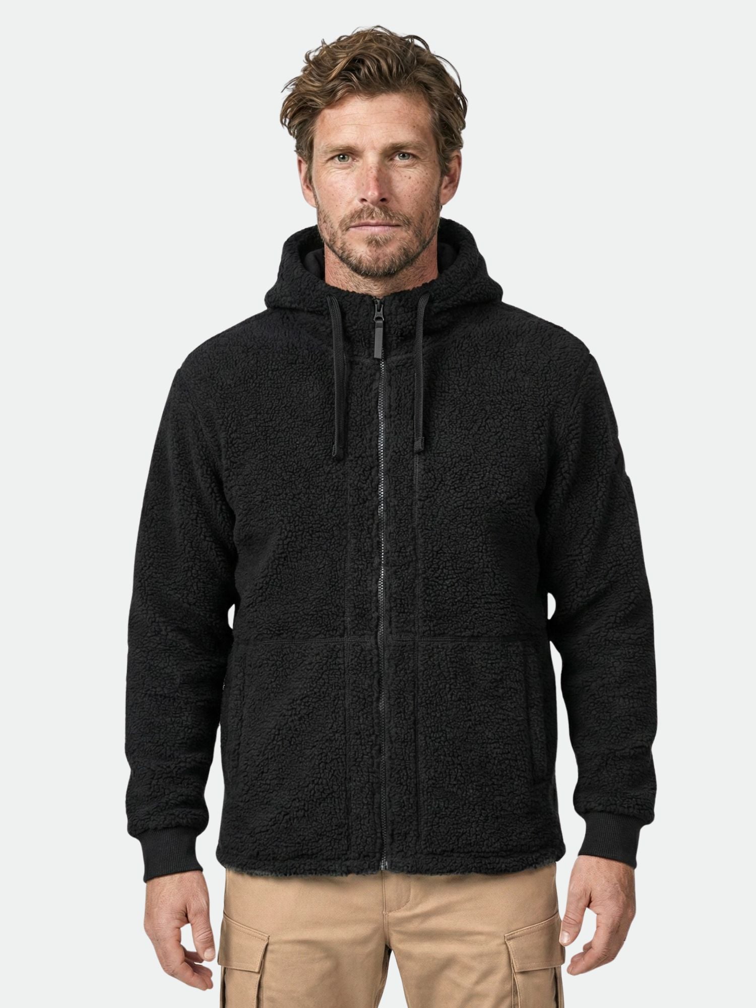 Ein Mann trägt einen schwarzen Halti Lykky Fleece Jacke Herren Kapuzenpulli und eine beige Cargohose und steht vor einem schlichten Hintergrund.