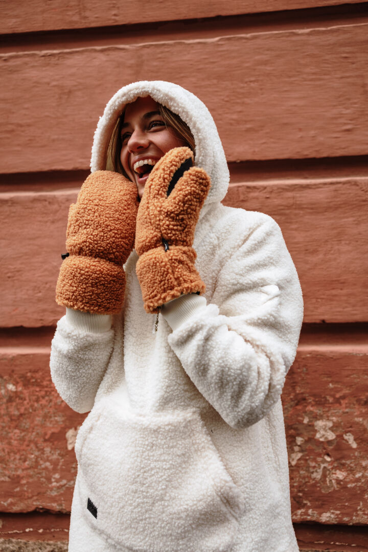 Person in einem weißen Halti Lykky Fleecepullover Damen und hellbraunen Fäustlingen lächelt vor einer strukturierten roten Wand.