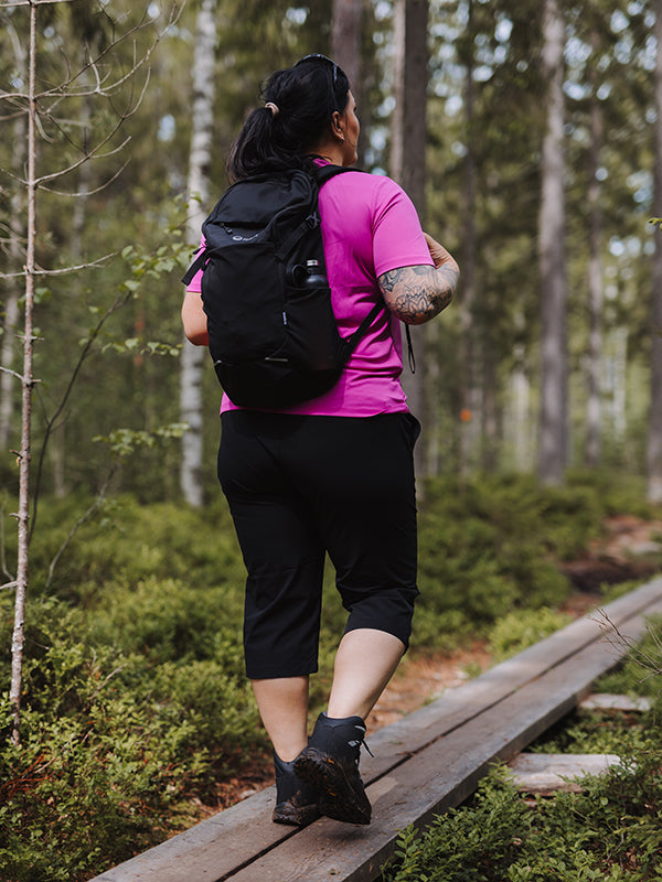 Person mit schwarzem Rucksack geht in Halti Drive X-Stretch Caprihose Damen Plus und rosa Shirt auf einem Waldweg.