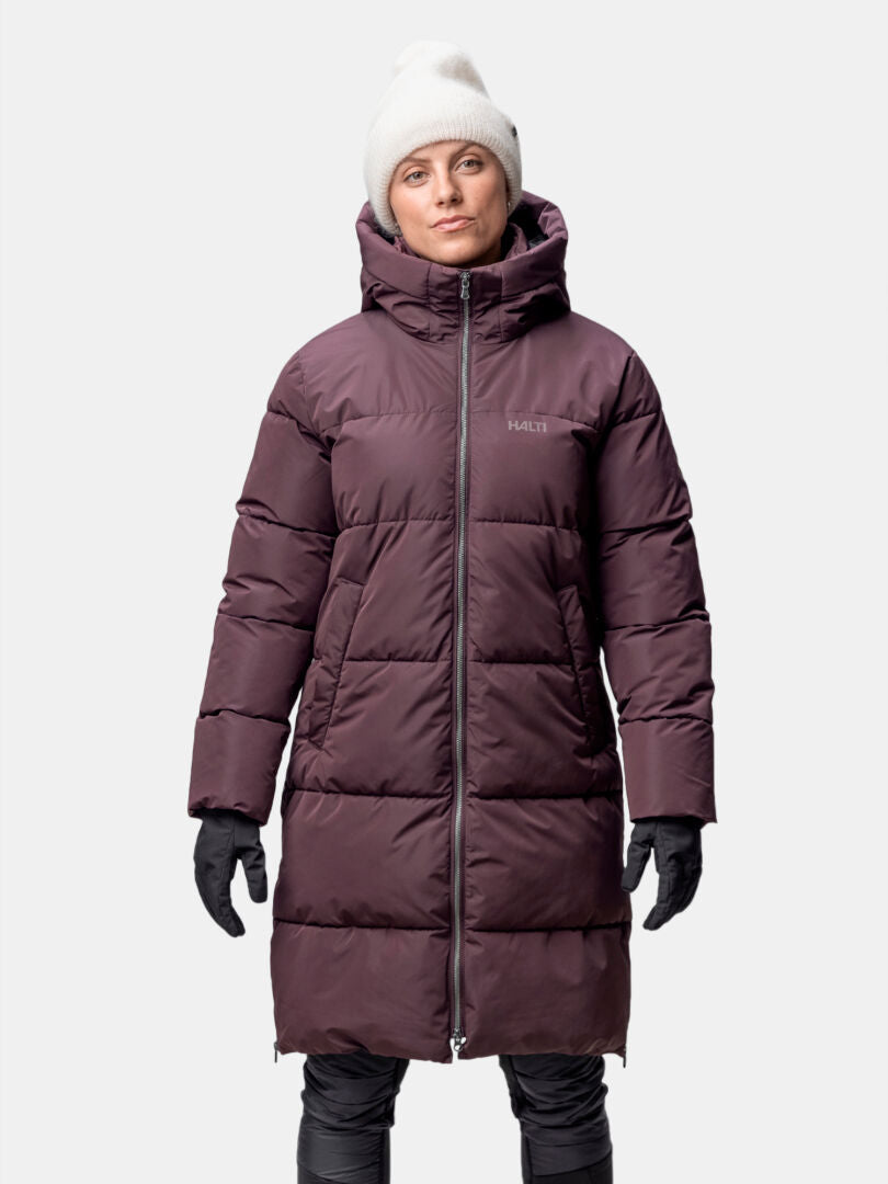 Eine Person steht mit dem Gesicht nach vorne in einer Halti Sammal Isolationsparka Damen, trägt schwarze Handschuhe und eine weiße Mütze.