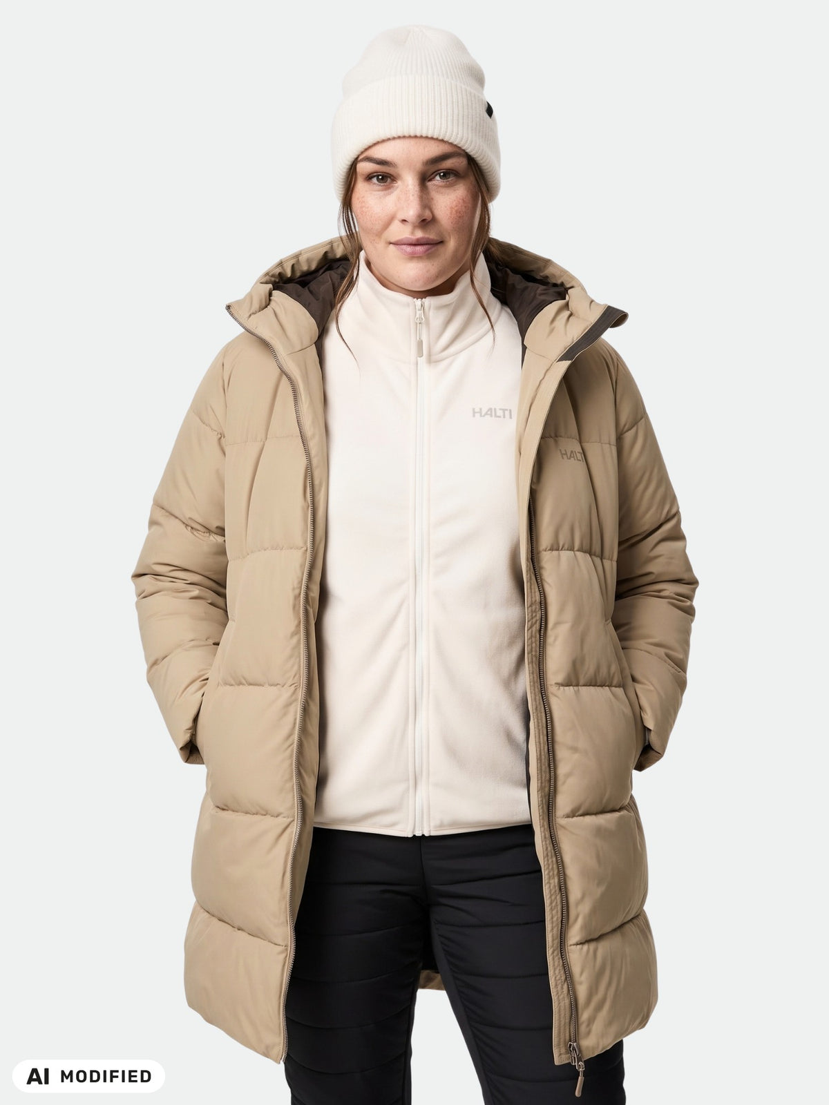 Frau in einem „Halti Sammal Isolationsparka Damen Plus“, einem weißen Oberteil mit Reißverschluss und einer Mütze, die Hände in den Taschen.