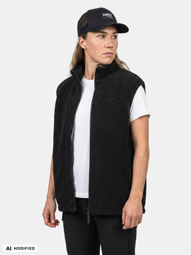 Person in einer „Halti Lykky Fleece Weste Unisex“, einem weißen T-Shirt und einer schwarzen Mütze vor einem einfarbigen, hellen Hintergrund.