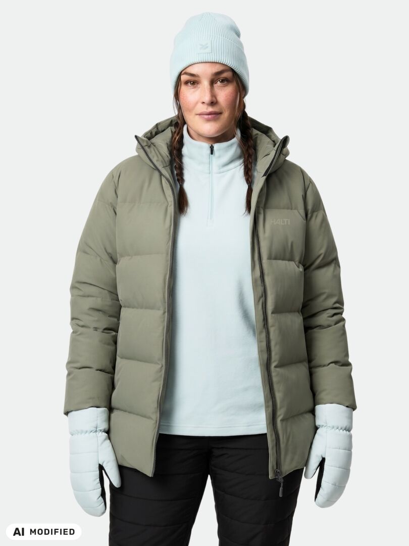 Eine Frau in einem grauen Halti Leinikko Puffer Parka Damen Plus, hellblauen Handschuhen und einer Mütze steht vor einem weißen Hintergrund.
