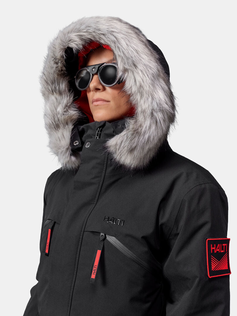 Person in Halti 1976 Down Parka Unisex, dunkler Sonnenbrille und roten Akzenten steht vor einem schlichten Hintergrund.