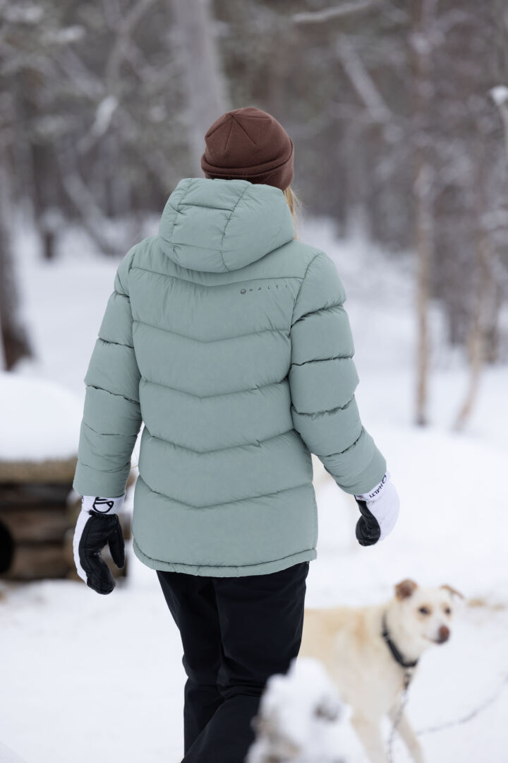 Eine Person in einem grünen Wintermantel und Halti Routa Winterhosen Damen geht neben einem Hund in einem verschneiten Wald spazieren.