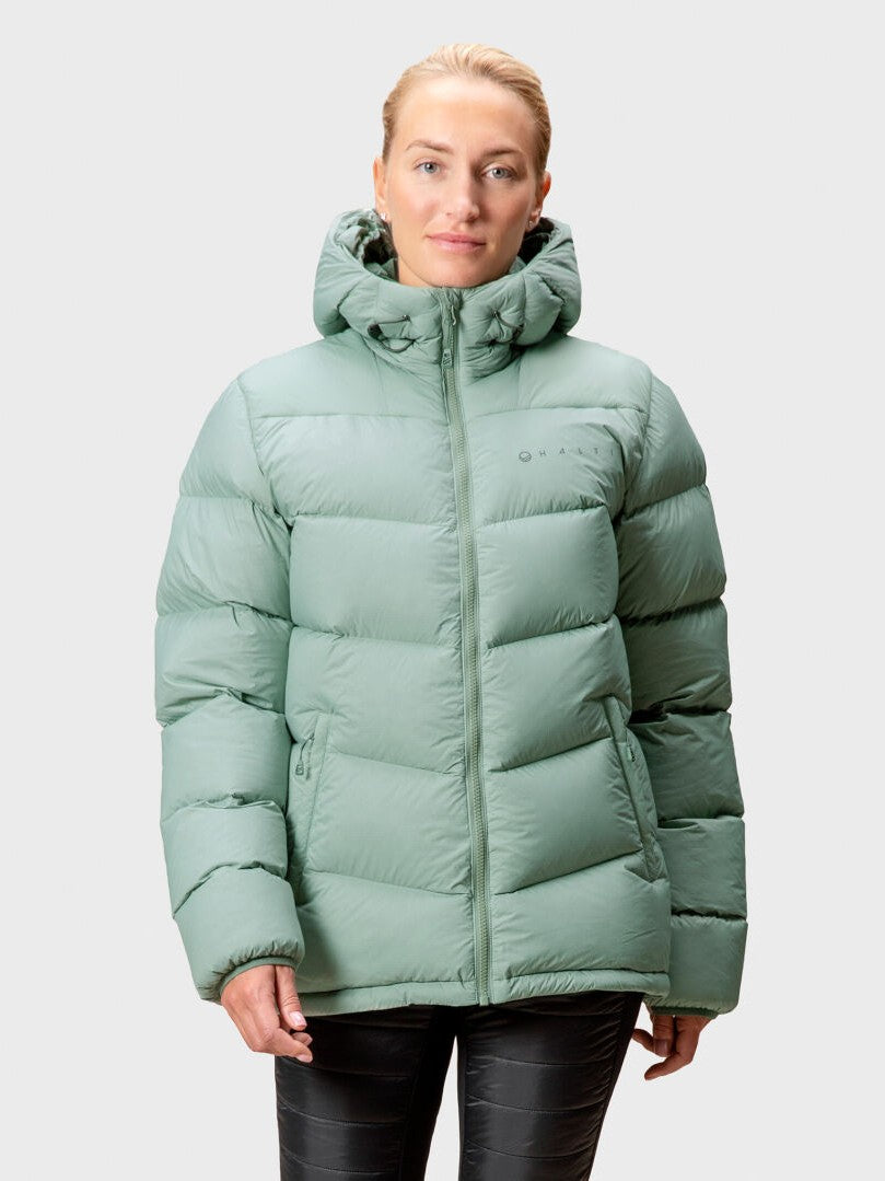 Eine Person in einer hellgrünen Halti Versant Daunenjacke Damen mit Kapuze, die vor einem schlichten hellen Hintergrund steht.