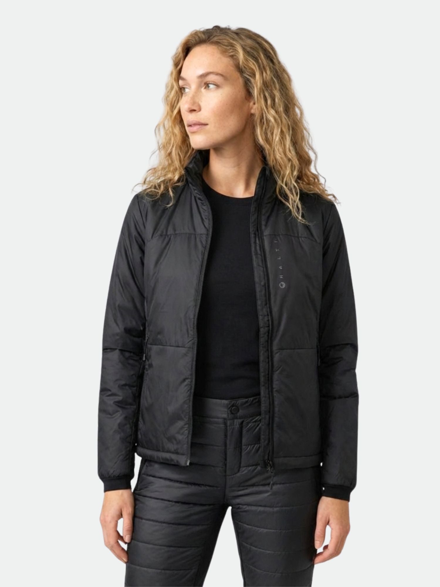 Eine Frau in einer schwarzen Halti Neon Primaloft Kunstfaserjacke Damen und Hose steht mit dem Gesicht nach links vor einem schlichten hellen Hintergrund.