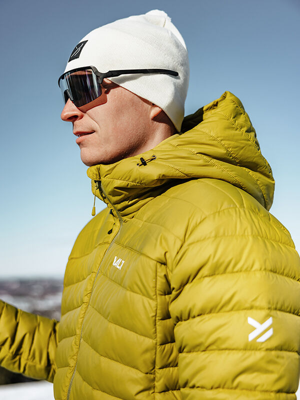 Eine Person in einer gelben Halti Huippu Daunenjacke Herren und weißer Mütze steht im Freien, bereit für sonnige Aktivitäten.