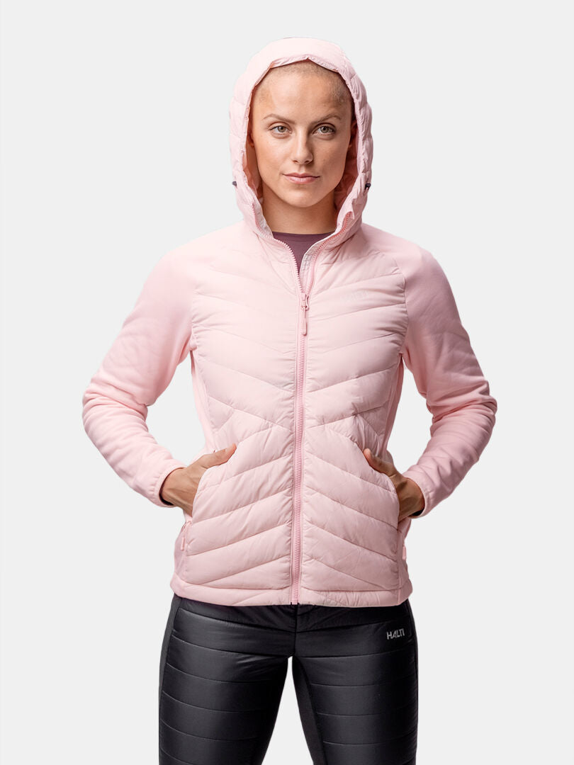 Person trägt eine hellrosa Halti Huippu Hybrid Daunenjacke Damen und schwarze Leggings, Hände in den Jackentaschen.