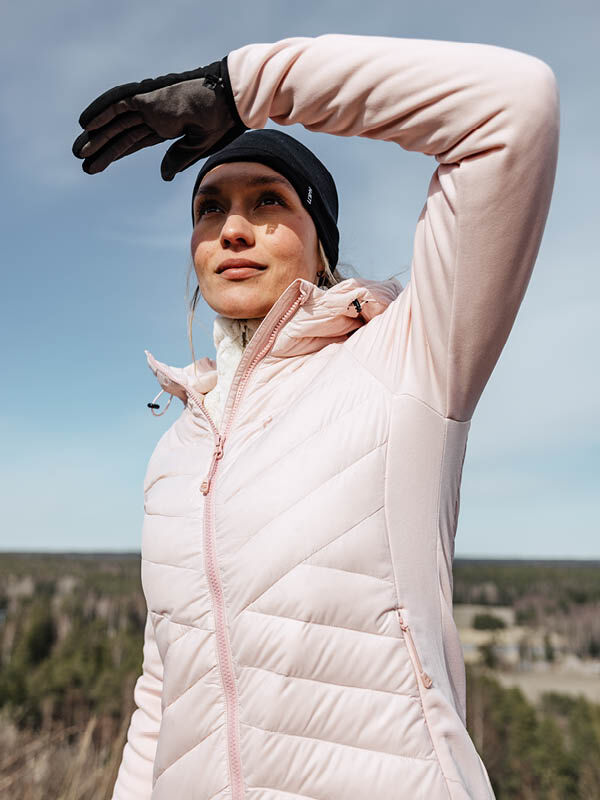 Eine Person in einem hellrosa Halti Huippu Hybrid Daunenjacke Damen und schwarzen Handschuhen schirmt ihre Augen vor der Sonne ab, dahinter Bäume.