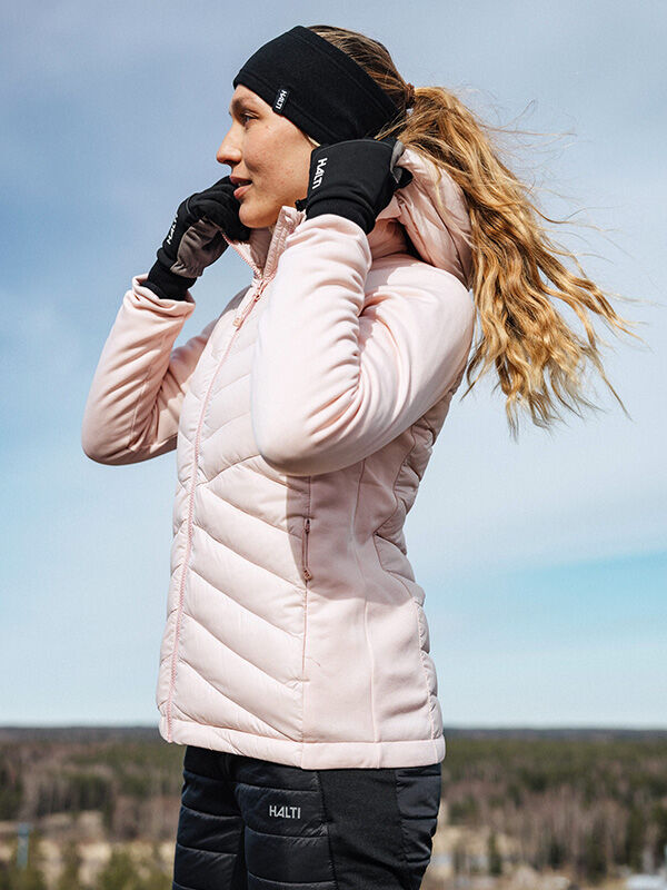 Eine Frau im Freien trägt eine Halti Huippu Hybrid Daunenjacke Damen, schwarze Handschuhe und ein Stirnband.