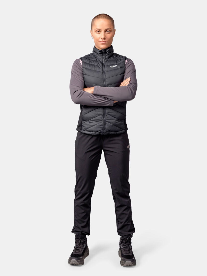 Eine Person blickt mit verschränkten Armen nach vorne und trägt ein Halti Huippu Hybrid Daunenweste Damen, langärmeliges Hemd, Hose und Stiefel.