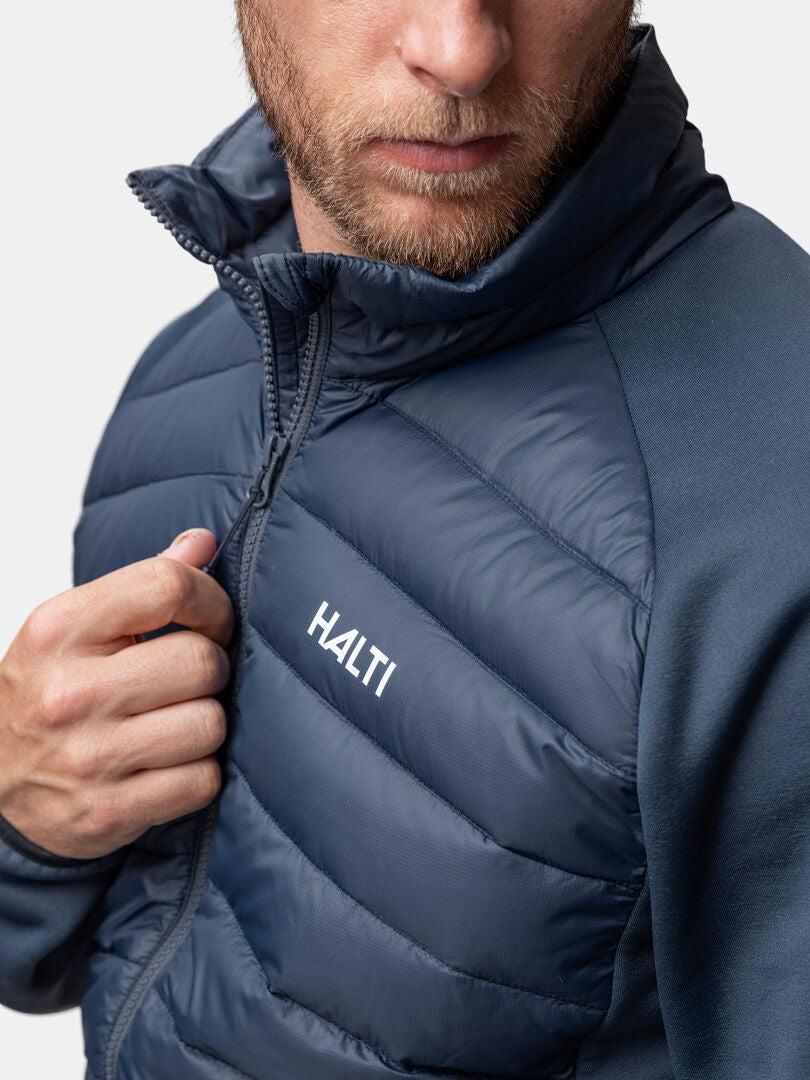 Ein Mann schließt den Reißverschluss einer Halti Huippu Hybrid Daunenjacke Herren, auf deren Brust das Logo der Marke zu sehen ist.