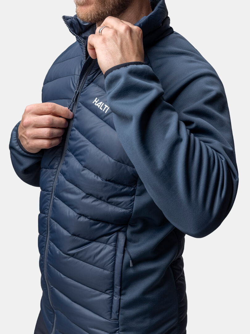 Ein Mann schließt den Reißverschluss einer blauen Halti Huippu Hybrid Daunenjacke, von den Schultern bis zur Mitte des Oberschenkels gesehen.