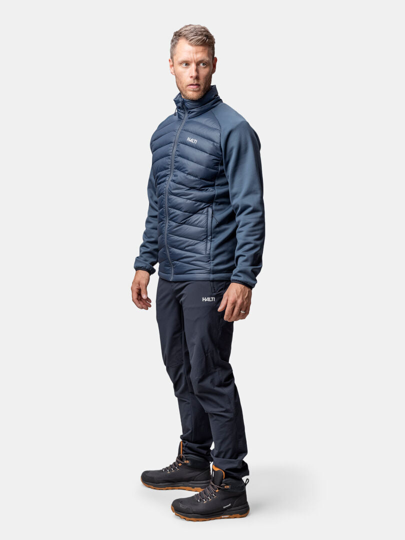 Ein Mann trägt die Halti Huippu Hybrid Daunenjacke Herren in navy mit passender Hose und schwarzen Stiefeln auf hellem Grund.