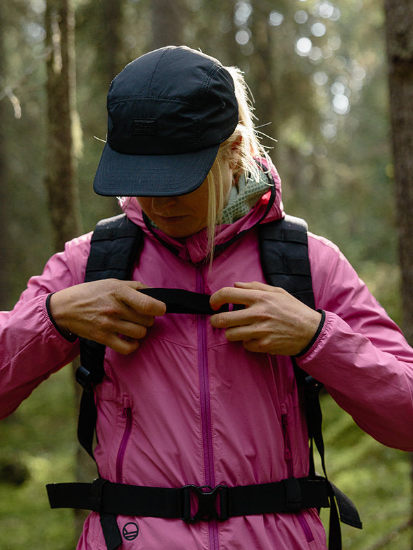 Eine Person, die eine rosafarbene Jacke und Halti Staffas Kappe trägt, stellt im Wald die Riemen ihres Rucksacks ein.