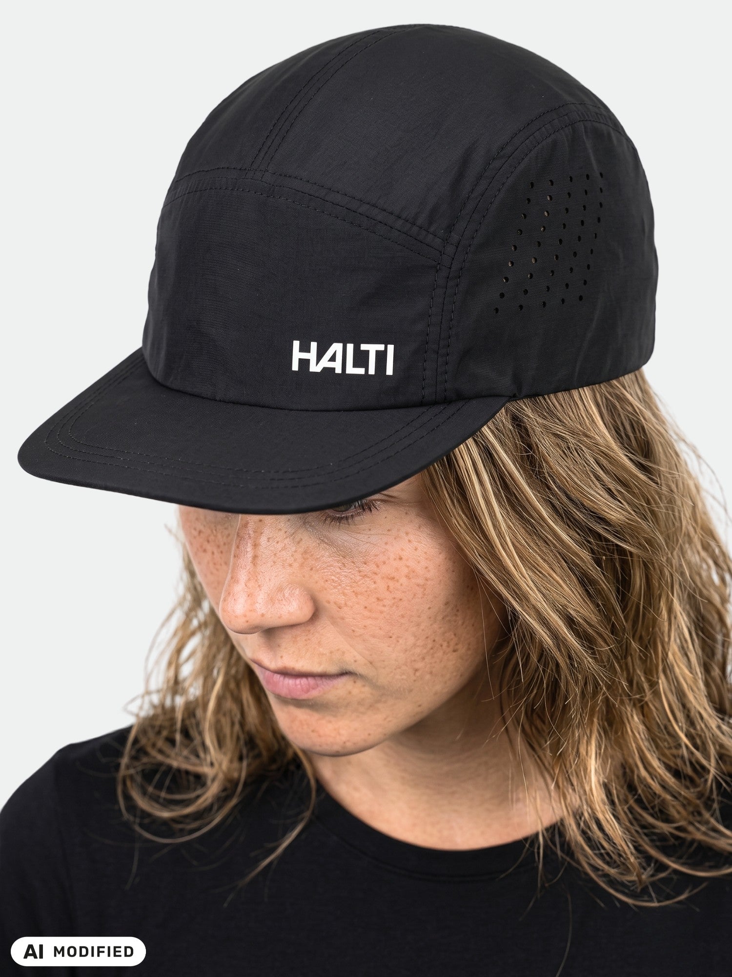 Eine Person mit gewelltem Haar, die eine Halti Luovi-Mütze und ein Hemd trägt und vor einem hellen Hintergrund nach unten blickt.