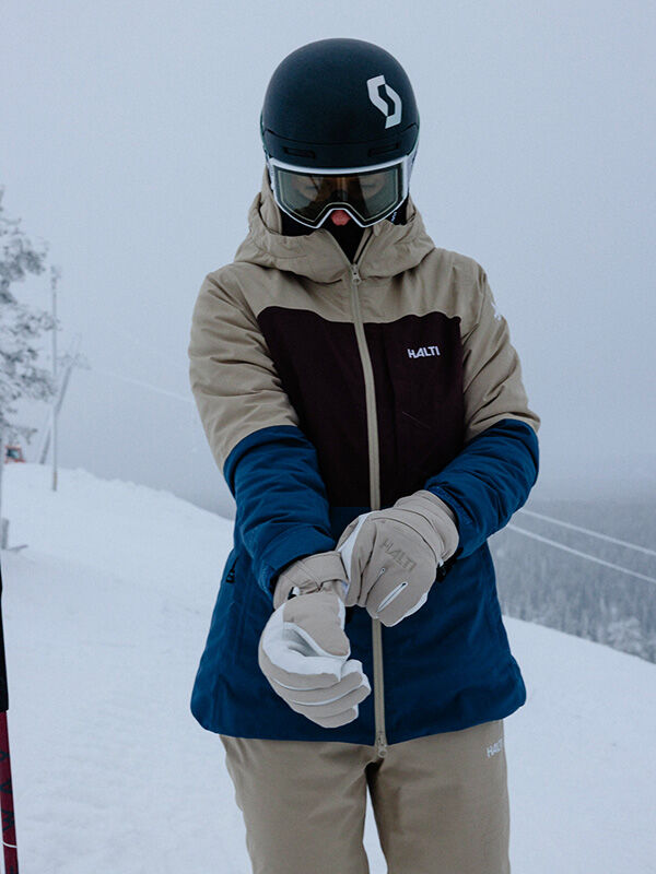 Ein Skifahrer mit Halti Vuotos Skihandschuhen und einem Helm steht auf einer verschneiten Piste und passt seinen Handschuh an.