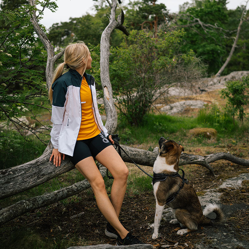 Eine Frau trägt die Halti Urbanite Damen Training Jacke und sitzt draußen auf einem Baumstamm, ihr Hund bei ihr.
