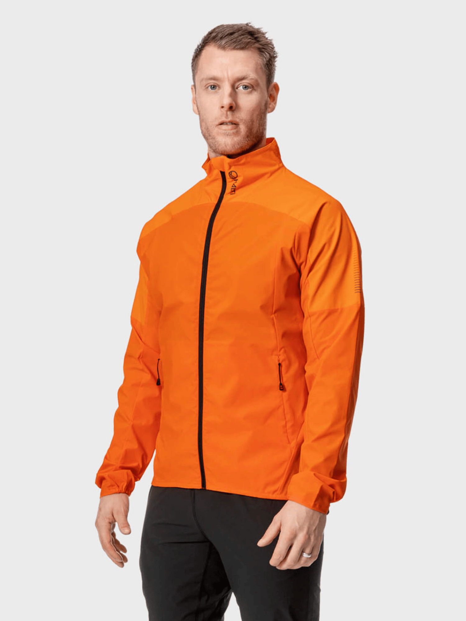 Ein Mann trägt die Halti Urbanite Herren-Trainingsjacke in leuchtendem Orange mit schwarzen Hosen und steht vor einem schlichten Hintergrund.