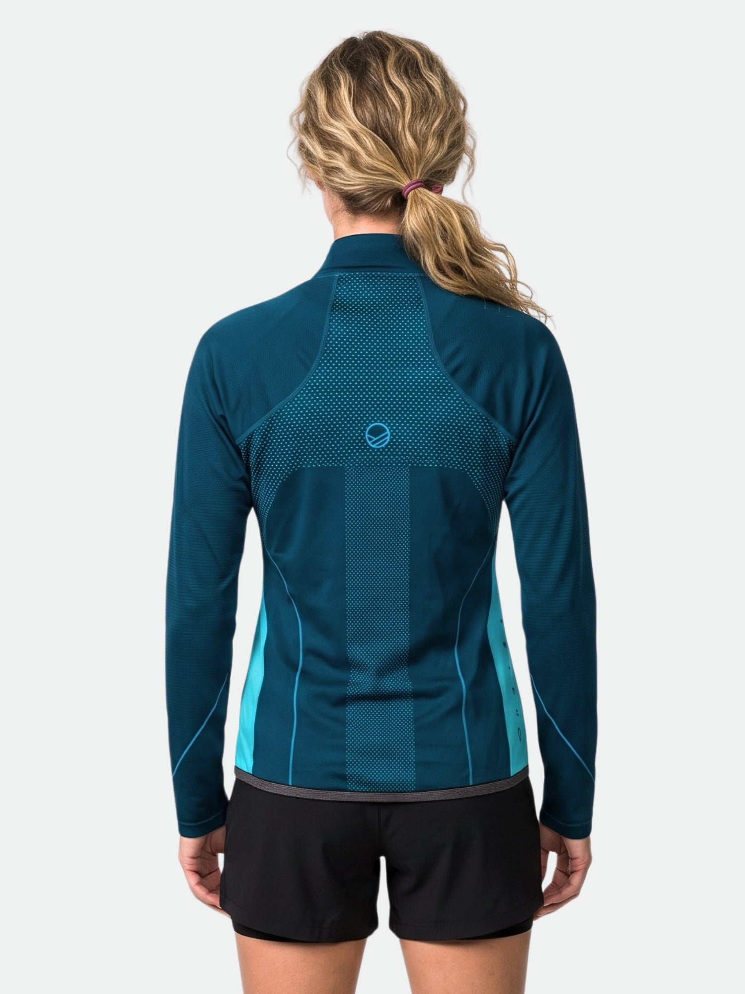 Eine Person mit welligem blondem Haar trägt eine „Halti Tracks Damen Seamless Jacke“ und schwarze Shorts; sie steht mit dem Rücken zur Kamera vor einem einfarbigen Hintergrund.