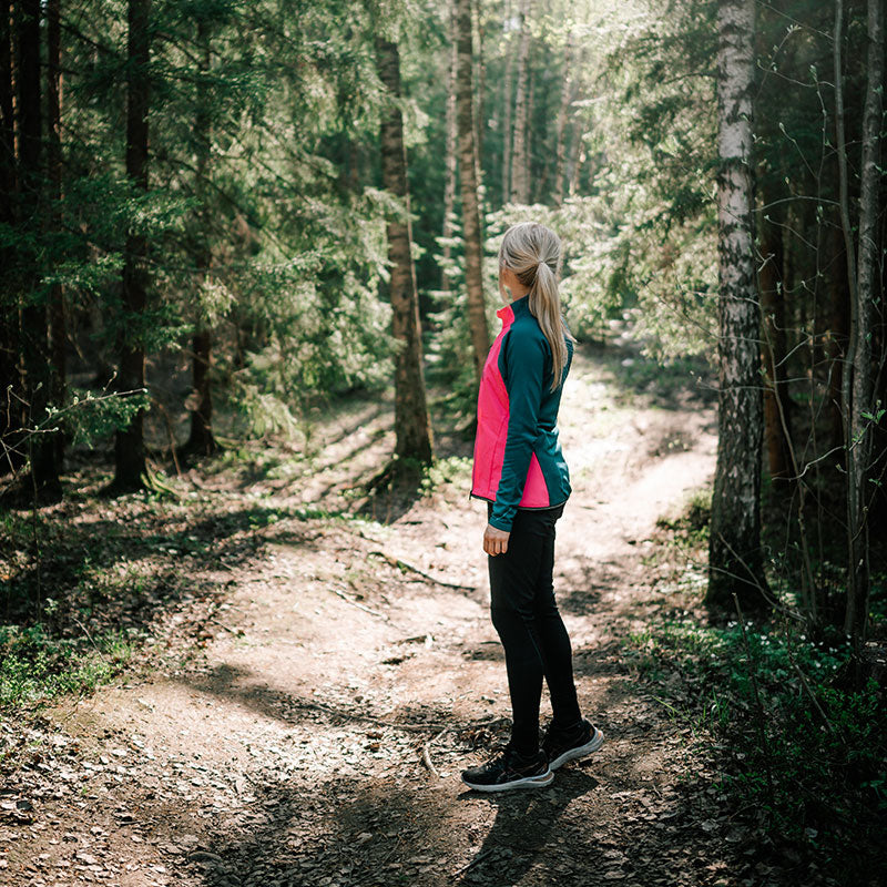 Eine blonde Frau in einer Halti Tracks Damen Seamless Jacke steht auf einem Feldweg in einem sonnigen Wald, bereit für Outdoor-Workouts.