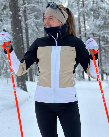 Eine Frau fährt in einem Wald Ski. Sie trägt die Halti Vinha Langlaufjacke Damen aus Stormwall®-Gewebe und hält orangefarbene Stöcke in der Hand.