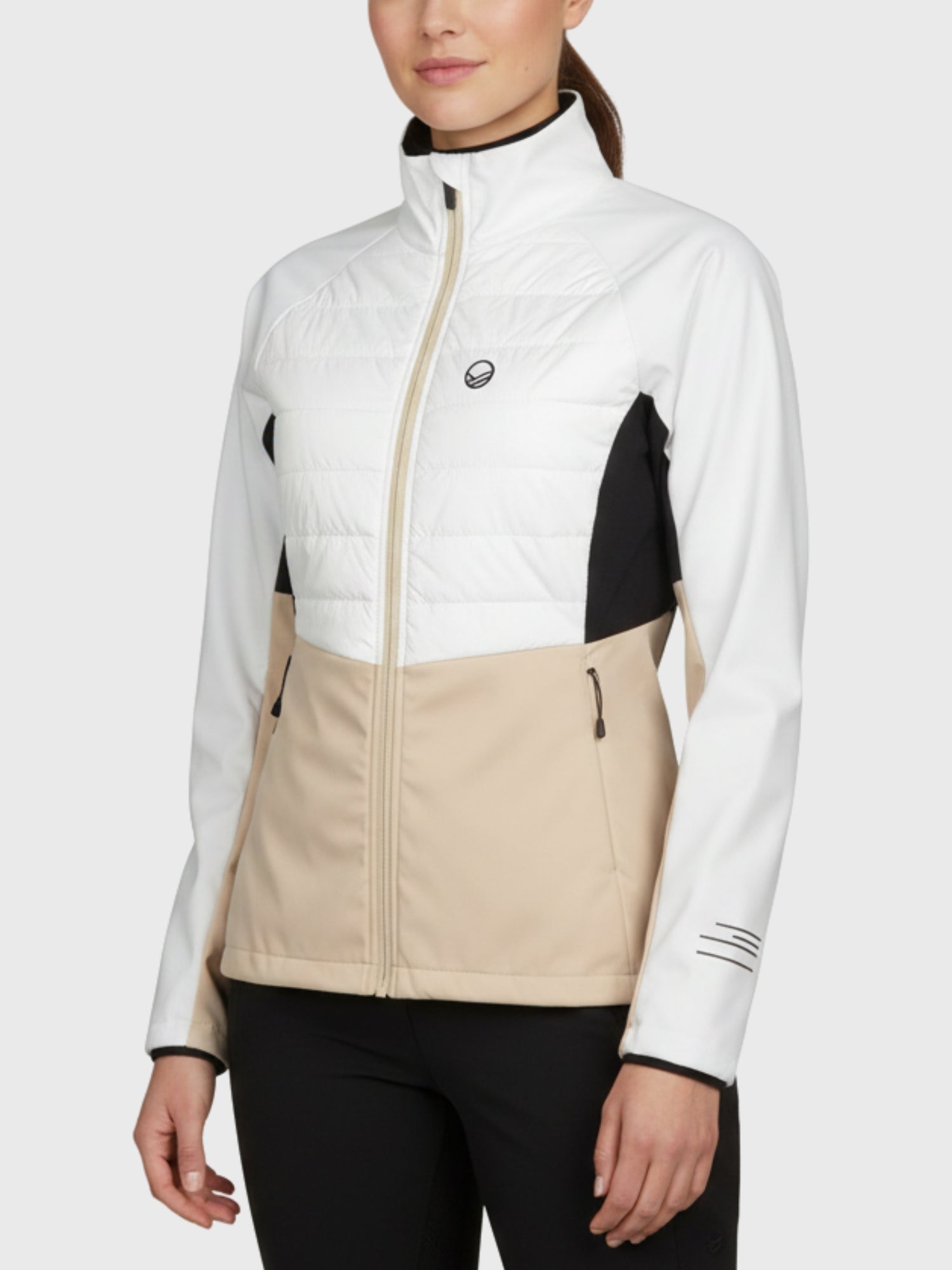 Eine Frau trägt die Halti Vinha Hybrid Langlaufjacke Damen in Weiß, Beige und Schwarz vor einem schlichten hellen Hintergrund.