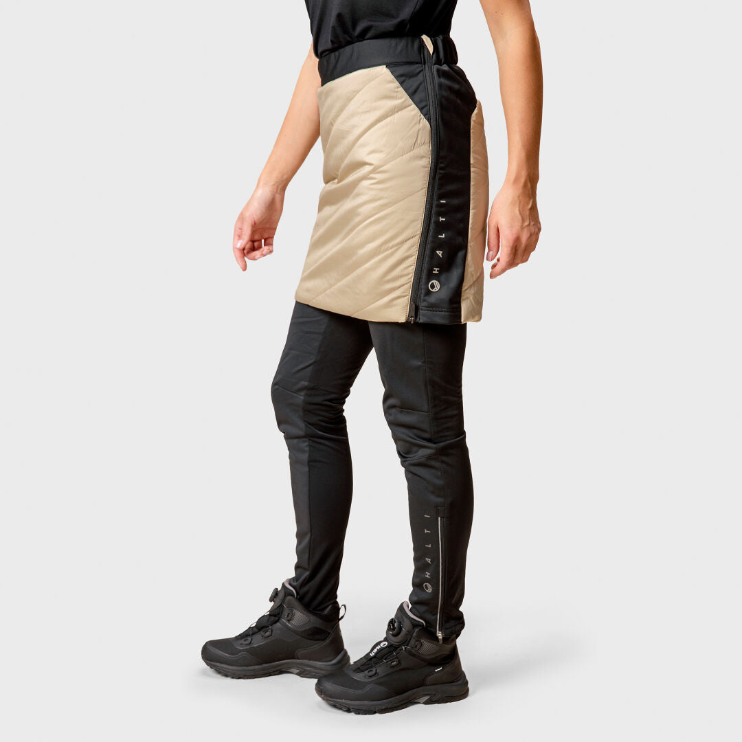 Person mit schwarzen Schuhen, engen Hosen und einem beigen Halti Hanki Hybrid Rock Damen, die vor einem schlichten Hintergrund steht.