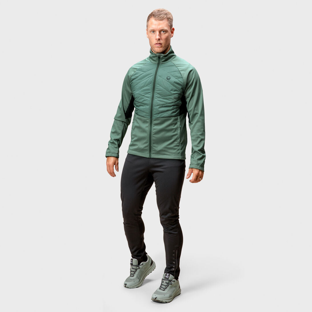 Ein Mann in einer grünen Halti Vinha Hybrid Langlaufjacke für Herren, schwarzen Hosen und grünen Schuhen steht vor einem schlichten Hintergrund.