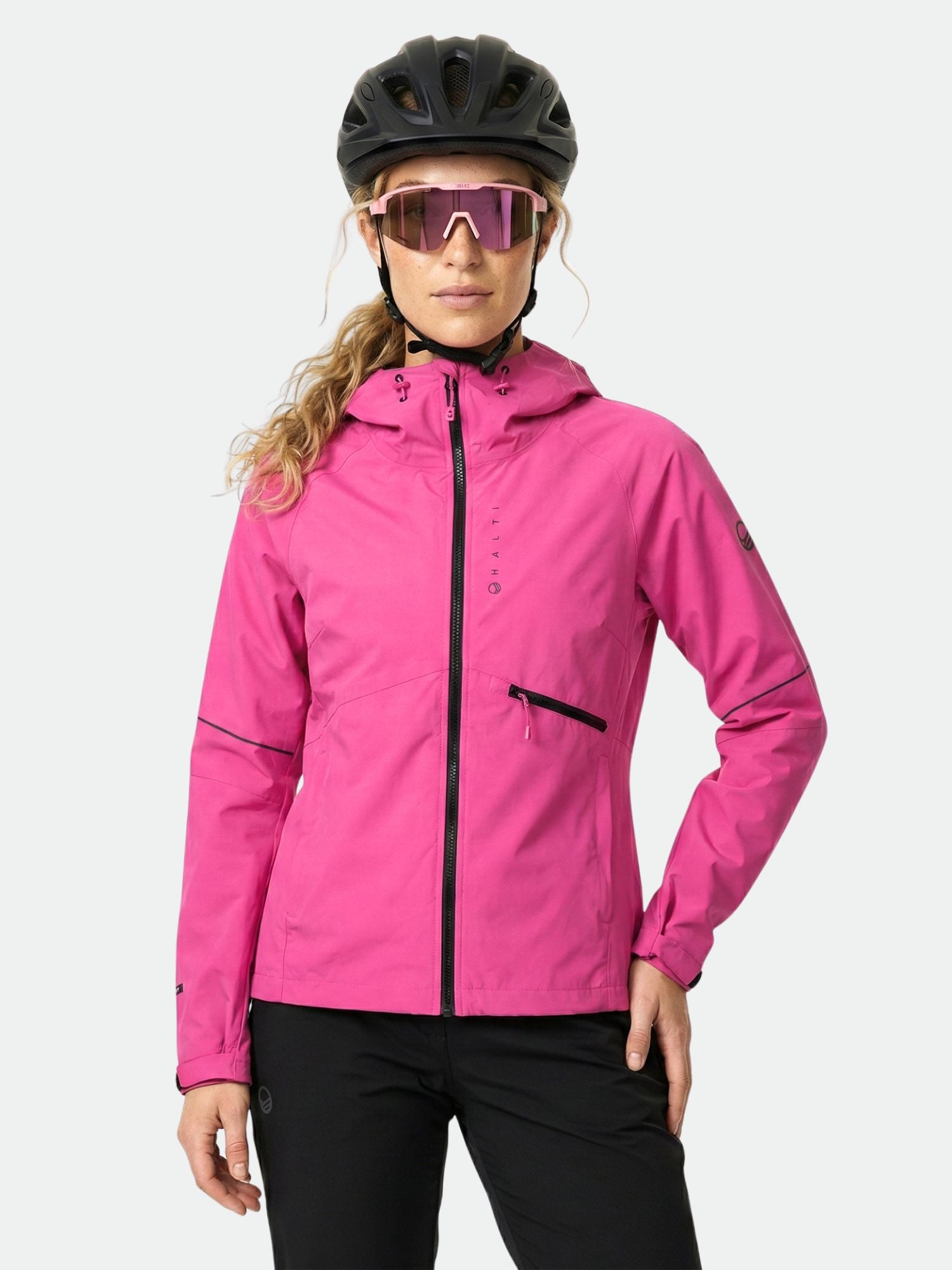 Eine Frau in einer pinkfarbenen Halti Shelter Stormwall 3L Jacke Damen, Helm und Sonnenbrille posiert selbstbewusst beim Winterradfahren.