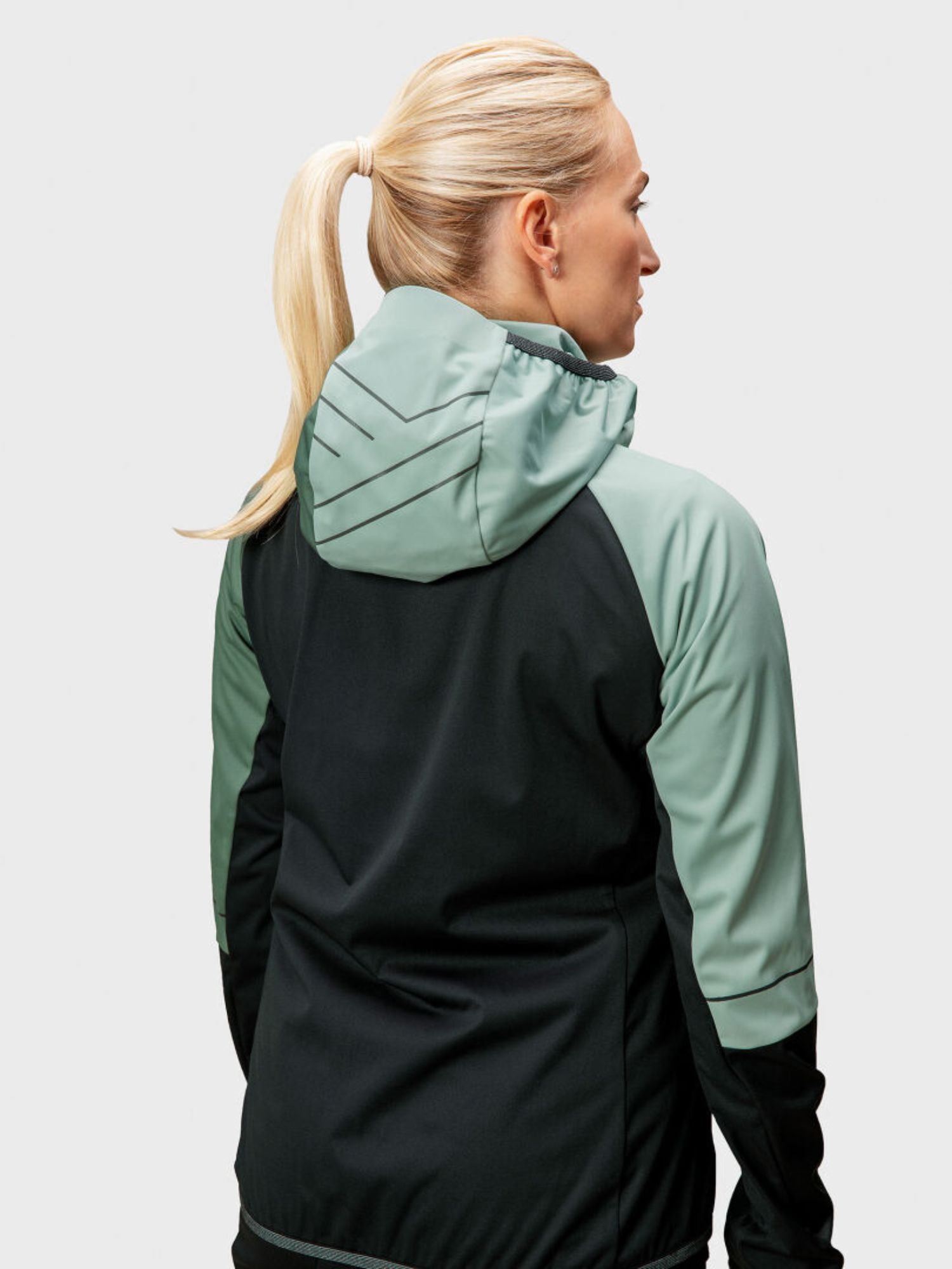 Eine blonde Frau mit Pferdeschwanz trägt die Halti Exhale Stormwall Jacke Damen mit reflektierenden Details, von hinten gesehen.