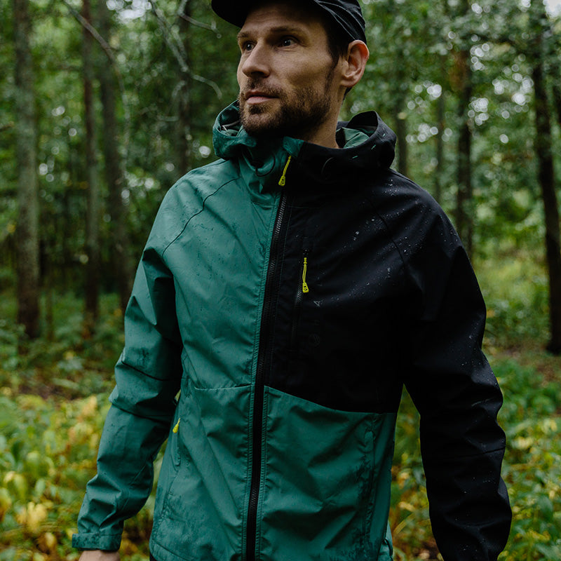 Ein Mann in einer Halti Shelter Stormwall 3L Jacke für Herren und einer schwarzen Mütze steht in einem Wald, bereit für Winteraktivitäten.