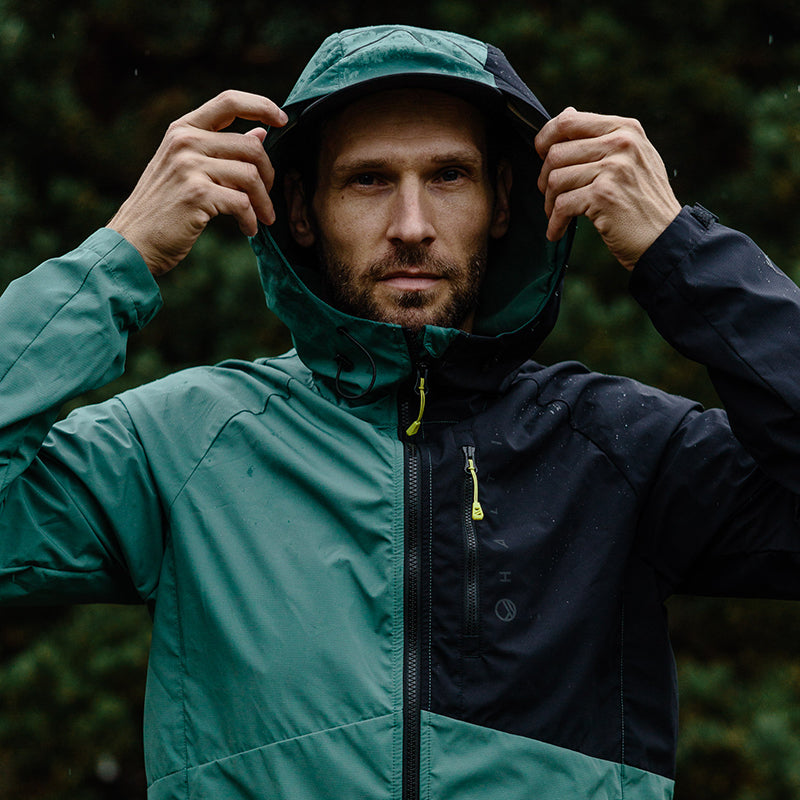 Ein Mann in einer Halti Shelter Stormwall 3L Jacke für Herren steht im Freien und hält die Ränder seiner Kapuze fest.