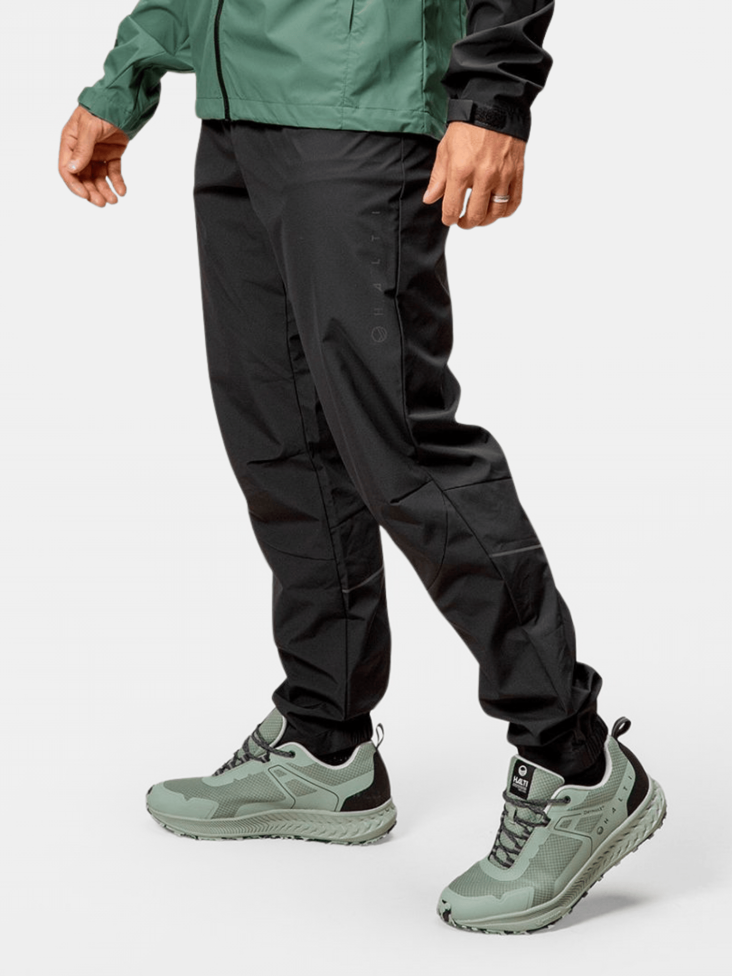 Eine Person trägt eine Halti Shelter Stormwall 3L Hose für Herren, eine grüne Jacke und hellgrüne Sportschuhe vor einem schlichten Hintergrund.