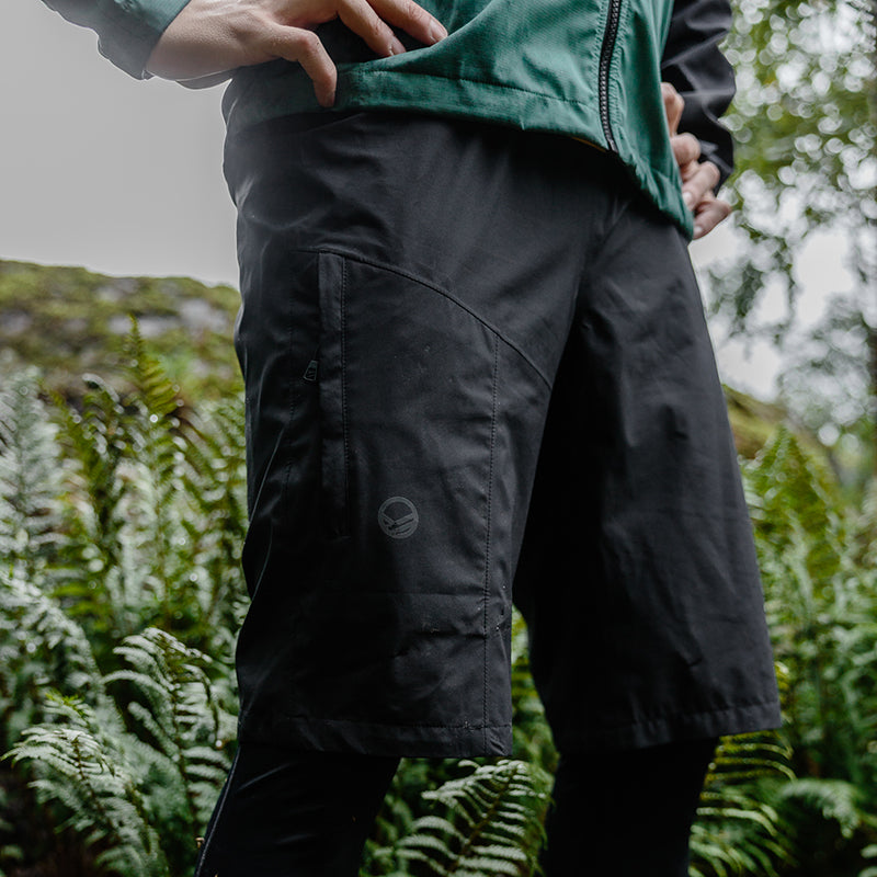 Person in grüner Jacke und Halti Shelter DrymaxX Shorts Unisex steht draußen zwischen Farnen, mit reflektierenden Details.