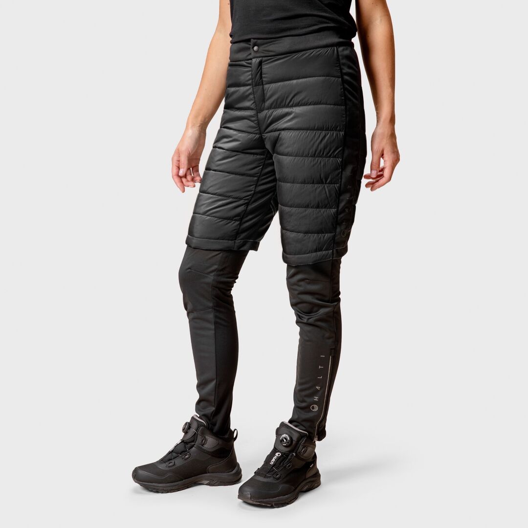 Die Halti Hanki Warm Hybrid Shorts Damen über Leggings mit Wanderschuhen, bereit für winterliche Aktivitäten vor einem schlichten Hintergrund.